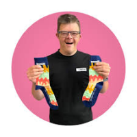 A smiling man with down syndrome on a purple pink circle background holds up the socks holds one in each hand 